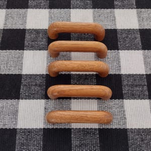 May include: Five matching cabinet pulls made of light brown wood with a visible natural grain. Each pull features a smooth, rounded arch shape with two mounting points on the underside, arranged in a vertical column on a checkered surface.