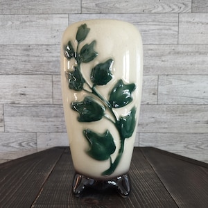 May include: A cream-colored ceramic vase featuring a raised, glossy dark green ivy leaf pattern on the front. The cylindrical vessel has a smooth, rounded body and is supported by three small, dark brown feet at the base, creating a stable stand.