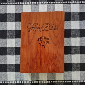 May include: A rectangular wooden box with a smooth, reddish-brown wood grain finish. The lid features the engraved text Holy Bible in a cursive script, positioned above a circular graphic containing a detailed illustration of a single rose.