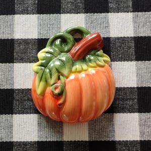May include: A ceramic pumpkin with a glossy finish. The pumpkin is orange with green leaves and a brown stem. The leaves have yellow highlights. The pumpkin is set against a black and white checkered background.
