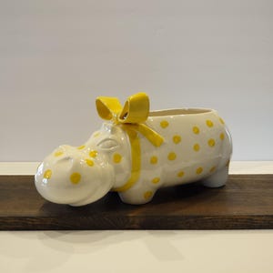May include: A ceramic planter shaped like a hippo, painted white with yellow polka dots. A yellow bow adorns the hippo's head. The planter sits on a dark wooden surface, with a white background.