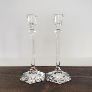May include: Two clear glass candlesticks with a tall, slender design. Each candlestick has a square base and a detailed top to hold a candle. The glass is transparent, reflecting light. The candlesticks are placed on a dark wooden surface.