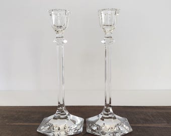 May include: A pair of clear glass candlesticks. They feature a tall, slender design with a square base, a long stem, and a small cup for a candle. The glass has a faceted look, reflecting light. The candlesticks are on a dark wooden surface. (approx. 30 cm tall)