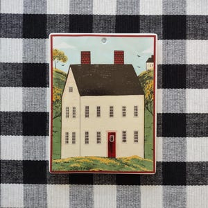 May include: A rectangular decorative tile featuring a quaint white house with a brown roof, red door, and chimneys. The house is set against a backdrop of green hills and a blue sky. The tile is placed on a black and white checkered tablecloth.