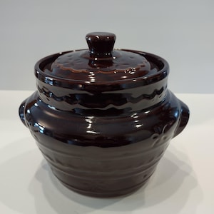 May include: A dark brown ceramic casserole dish with a lid. The dish has two handles and a decorative pattern around the rim and on the lid. The lid has a small knob for easy handling.