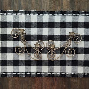 May include: Two silver-toned candle holders with ornate, swirling designs. Each holder has three candle cups and rests on a black and white checkered table runner. The runner is placed on a dark wooden surface.