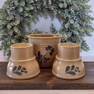 May include: A set of three tan ceramic vessels with a glossy finish. The central vessel is a tall, wide-mouthed pot featuring a dark blue painted flower design. Two smaller, matching vessels with flared bases and dark blue leaf motifs sit in front.
