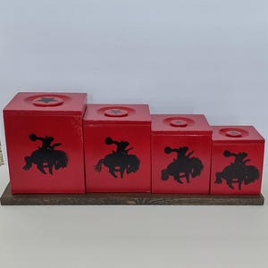 May include: Four red wooden boxes of varying sizes, arranged in a stepped configuration on a wooden base. Each box features a black silhouette of a cowboy riding a horse. The tops of the boxes have a star design.