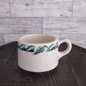 May include: A white ceramic mug with a dark green wave and leaf pattern around the rim. The mug has a rounded handle and a cylindrical shape. The mug is sitting on a dark wooden surface.