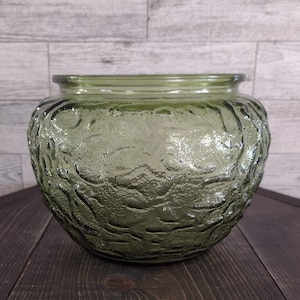 May include: A round, olive green glass bowl with a textured, organic pattern. The bowl has a wide opening and a slightly flared rim. The glass has a subtle, translucent quality, and the bowl sits on a dark wooden surface.