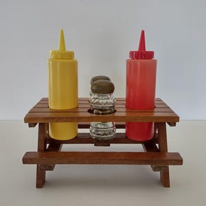 May include: A miniature wooden picnic table condiment holder featuring a yellow squeeze bottle on the left, a red squeeze bottle on the right, and a clear glass salt or pepper shaker with a brown lid positioned in the center of the table.