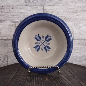 May include: A ceramic bowl with a white interior and a dark blue rim. The bowl features a blue floral design in the center. The bowl is displayed on a silver metal stand against a wooden background.