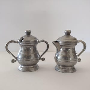 May include: Two antique pewter serving pieces. One is a small creamer with a spout and handle. The other is a sugar bowl with a lid, handle, and a slot for a spoon. Both pieces have a textured finish and are on a white background.
