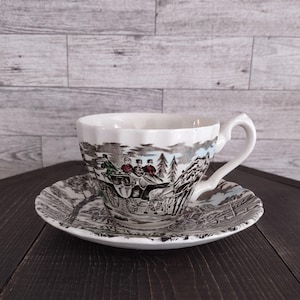 May include: White teacup and saucer set with a black and white transferware design depicting a horse-drawn carriage scene. The cup has a scalloped rim and a curved handle. The saucer mirrors the cup's design.