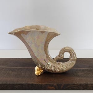 May include: A vintage, iridescent ceramic cornucopia vase. The vase is a creamy, pearlescent color with gold accents on the base and rim. The horn-shaped vase has a curved handle and a ruffled edge.