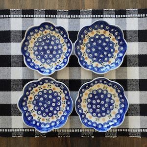 May include: Four scalloped ceramic plates featuring a cream-colored base with a repeating pattern of blue circles and small green and yellow floral accents. Each plate has a distinct wavy edge and a central cluster of blue circular motifs.