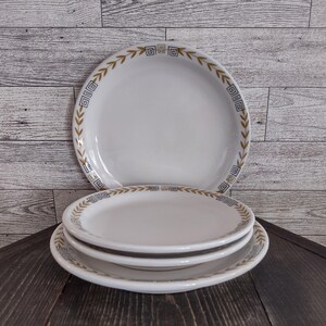 May include: A set of three white ceramic plates stacked vertically. Each plate features a decorative border consisting of a repeating gold leaf pattern interspersed with small blue and gold square geometric Greek key motifs along the rim.