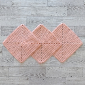 May include: Three square crochet dishcloths in a soft peach color. Each piece features a diamond-shaped pattern created with a central cross of openwork stitches, radiating outward from the center to the edges in a consistent texture.