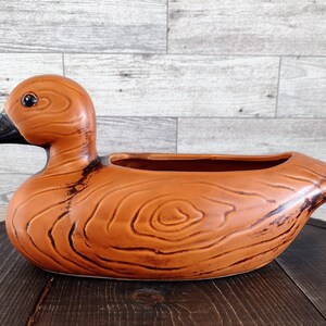 May include: A ceramic duck-shaped planter featuring a glossy orange-brown finish with dark brown wood grain patterns. The duck has a smooth, rounded body, a black beak, and a single black eye. The top of the duck's back is open for planting.