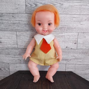May include: A vintage doll with orange hair, wearing a yellow romper with a white collar and a red tie. The doll has a smiling expression and is posed on a dark surface.