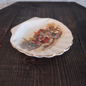 May include: A white ceramic shell-shaped dish with a gold-trimmed edge. The center features a detailed, colorful illustration of figures in historical attire standing near a building and a carriage. The text "Dr. Johnson" is printed below the scene.