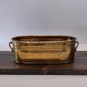 May include: An oval-shaped brass planter with a polished metallic finish, featuring horizontal ribbed detailing along the sides and two small, curved handles on opposite ends. The container has a wide, open top and a flat base for stability.
