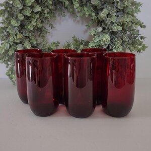 May include: A set of six cylindrical drinking glasses in a deep, translucent ruby red color. The glasses feature a smooth, seamless design with rounded bases and straight sides, arranged in a cluster to display their uniform shape and rich color.