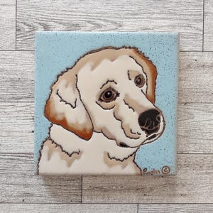May include: Square ceramic tile featuring a cream-colored dog portrait with brown accents, set against a light blue speckled background. The dog has brown eyes and a black nose. The tile is signed "Pumpkin".