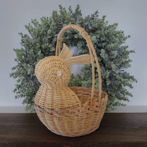 May include: A light beige woven wicker basket shaped like a rabbit with a single tall arched handle. The basket features a rounded body, a prominent head with two long ears, and a small white circular eye detail on the side of the rabbit head.