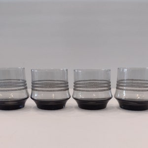 May include: Four clear drinking glasses with a black base and three horizontal gray bands. The glasses are cylindrical with a slightly flared base. The gray bands have a textured appearance. The glasses are arranged in a row.