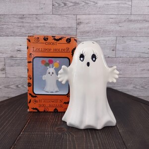 May include: A white ceramic ghost figurine with painted black eyes and a small mouth, standing next to its orange and black cardboard box labeled Ghost Lollipop Holder and Halloween Boo Bazaar, featuring a graphic of the ghost holding lollipops.