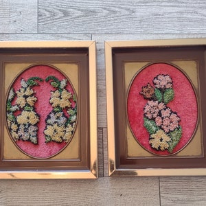 May include: Two framed floral needlepoint artworks. Each piece features an oval design with a pink background and a gold frame. The needlepoint depicts colorful flowers in shades of yellow, green, and pink.