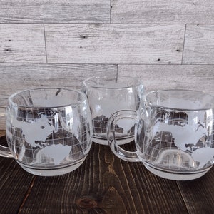 May include: Three clear glass mugs featuring a frosted white etched world map design with longitudinal and latitudinal grid lines. Each mug has a rounded body, a sturdy handle, and a smooth rim, showcasing the continents of North and South America.
