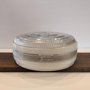 May include: A vintage, round glass light fixture with a clear, ribbed top and a white, frosted bottom section. The fixture has a decorative band with a geometric pattern. It is sitting on a dark wooden surface.