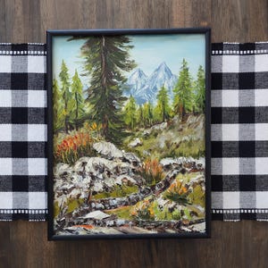 May include: An oil painting in a black frame depicts a mountain landscape. The artwork features a forest of green trees, rocky terrain, and snow-capped mountains under a blue sky. The painting is set against a black and white checkered tablecloth.