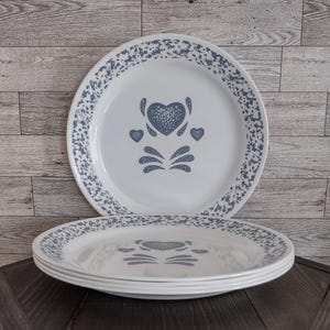 May include: A set of white dinner plates featuring a blue speckled rim pattern and a central blue heart motif surrounded by smaller heart and petal-shaped accents. The plates have a smooth, glossy finish and a consistent circular design.