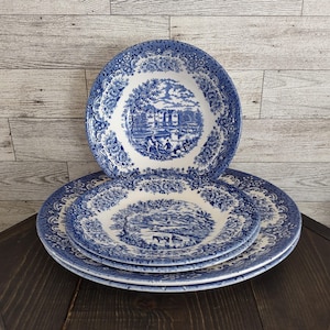 May include: A stack of three round ceramic plates featuring a blue and white transferware pattern. Each plate displays a central landscape scene with buildings and figures, surrounded by a wide border of intricate floral and leaf motifs.