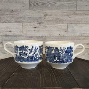 May include: Two white ceramic mugs with blue Willow pattern scenes. One mug features a boat, birds, and buildings, while the other depicts a pagoda and trees. Both mugs have a curved handle.
