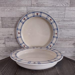May include: Two round ceramic dishes with a cream-colored center and a wide rim featuring a blue grid pattern. Small red heart motifs are placed within the grid squares along the border of each dish. The dishes have a smooth, glossy finish.