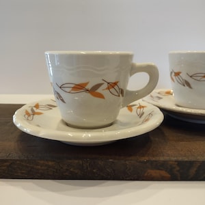 May include: A white ceramic teacup and saucer set featuring a repeating pattern of stylized orange and brown leaves. The cup has a curved handle and a subtle wavy embossed rim, while the matching saucer displays the same leaf motif.