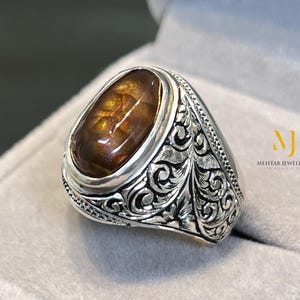 Könnte beinhalten: Ein silberner Ring mit einem ovalen, braunen Edelstein. Der Ring hat ein verziertes, graviertes Design um das Band. Der Edelstein ist in einer Zargenfassung gefasst. Der Ring wird in einer grauen Ringschatulle präsentiert. Der Text "MEHTAD JEWELLERS" ist sichtbar.
