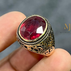 Può includere: Anello dorato con una gemma rosso scuro ovale e sfaccettata. L'anello presenta un ornato design a filigrana sulla fascia e intorno alla gemma. Il testo "MJ MOHAMED JEWELS" è visibile sul lato destro dell'immagine.