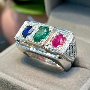 May include: A silver ring featuring three oval gemstones: blue, green, and red. The ring has a rectangular top with a detailed border and a textured side design. The ring is displayed in a gray velvet box.