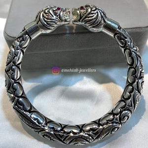 Puede incluir: Pulsera de plata con dos cabezas de león en los extremos. Las cabezas de león tienen ojos de piedras preciosas rojas. La pulsera está decorada con intrincados diseños florales.