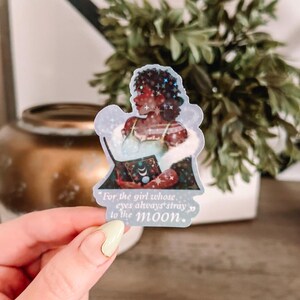May include: A sticker featuring a celestial illustration of a person holding a book. The person is depicted with dark skin, curly hair, and adorned with stars. The sticker includes the quote, "For the girl whose eyes always stray to the moon."
