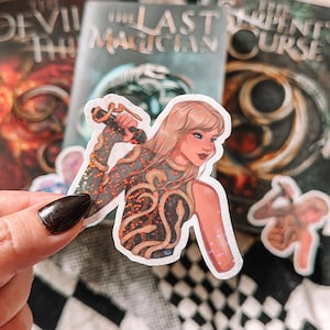 May include: A sticker featuring a woman with long blonde hair wearing a sparkly orange and black top with a snake design. The sticker is on a background of three book covers with the titles "The Devil's Thief", "The Last Magician", and "The Serpent's Curse".