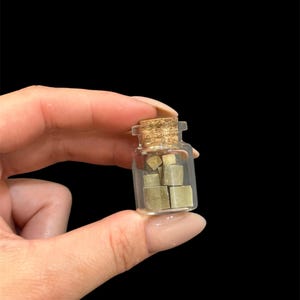 May include: A small, clear glass bottle with a cork stopper, filled with several small, square, light green objects. The bottle is held against a black background.