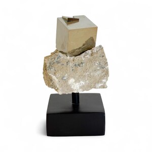 May include: A pyrite sculpture featuring a golden cube and a small gold triangle atop a rough, beige stone. The stone rests on a black stand. The sculpture is a unique decorative item.