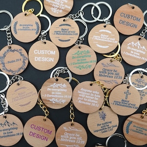 May include: A selection of wooden keyrings with metal rings. Each keyring has engraved text, including phrases like "Custom Design," Bible verses, and decorative elements. The keyrings have gold and silver-coloured rings.