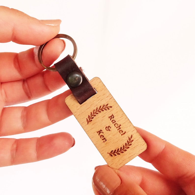 Op de afbeelding: Een houten sleutelhanger met een bruin leren bandje en een metalen ring. De sleutelhanger is gegraveerd met de namen "Rachel & Ken" en een laurierkransontwerp.
